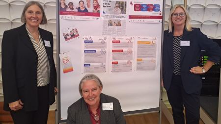 Drei Frauen stehen an einer Pinnwand mit Informationen zum Projekt "MY TURN. MY CAREER." und lächeln in die Kamera