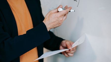 Eine Person steht vor einem Whiteboard. Es ist nur der Rumpf der Person und beide Arme zu sehen. Diese trägt ein gelbes Oberteil und darüber ein schwarzes Jackett. In der linken Hand hält sie ein Blatt Papier, in der rechten Hand einen Stift. 