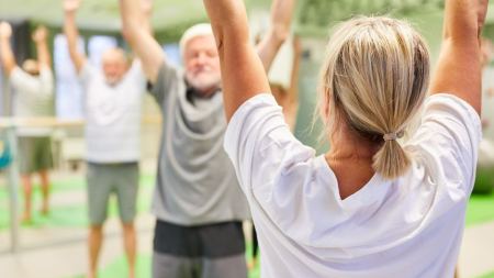Im Vordergrund steht eine Person mit weißem T-Shirt in Rückenansicht und hebt die Arme nach oben. Sie blickt ihr zugewandte Personen im Hintergrund an, die ebenfalls die Arme in die Höhe strecken.