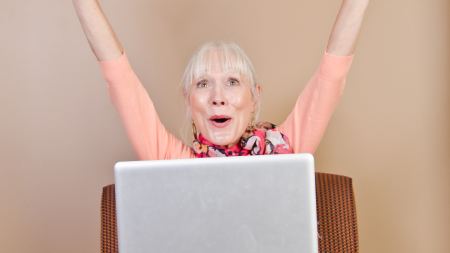 Ältere Frau sitzt auf Stuhl mit Laptop auf dem Schoß und jubelt 
