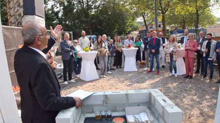 Mann mit Anzug steht mit dem Rücken zur Kamera, neben ihm ein Rednerpult, vor ihm ist in der Ferne eine Menschengruppe zu sehen, die an Stehtischen stehen und den Mann anschauen