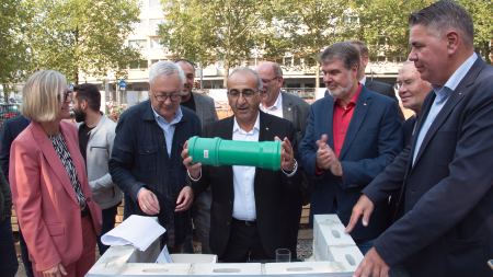 Eine Gruppe feierlich gekleideter Menschen steht hinter einem Podest mit der Aufschrift "Grundsteinlegung", der Mann in der Mitte hält ein Rohr in beiden Händen