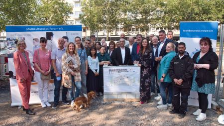 Eine Gruppe feierlich gekleideter Menschen steht vor zwei Bannern vor einer Baustelle, in der Mitte befindet sich ein Rednerpult