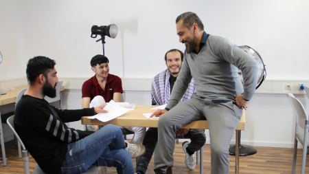 Drei Lernende sitzen um einen Tisch, ein Dozent lehnt an den Tisch und zeigt auf ein Dokument