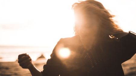 Eine Person mit langen Haaren verdeckt die Sonne. Sie fasst sich mit einer Hand ins Haar. Im Hintergrund ist Sand und Meer zu erkennen.