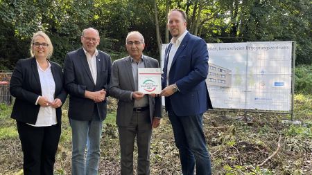 Übergabe Förderbescheid Neubau Stiftung Eine Frau und drei Männer stehen auf einem Baugrundstück, einer der Männer hält eine Plakette in der Hand