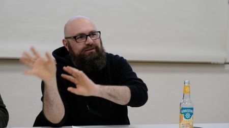 Sascha Schiessl (Mann mit Glatze, Bart und Brille) sitzt an einem Tisch und hält die Unterarme hoch. Vor ihm steht ein Tisch mit Unterlagen und einer Getränkeflasche darauf. 