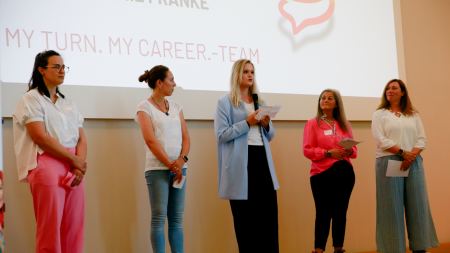 Fünf Frauen stehen nebeneinander auf der Bühne vor einer Leinwand. Eine Frau hält ein Mikrofon in der Hand.