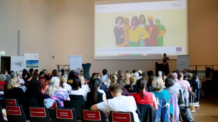 Aufnahme mit Rückenansicht eines Publikums in einem großen Saal. Ganz vorne wird eine Beamerprojektion an die Wand geworfen.