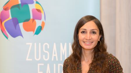 Gilda Sahebi steht vor einem Banner mit einem Globus-Motiv aus bunten Sprechblasen - darunter sieht man Teile des Schriftzugs Zusammen Fair.