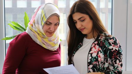 zwei junge Frauen - links mit Kopftuch, rechts mit langen braunen Haaren - schauen auf einen Zettel