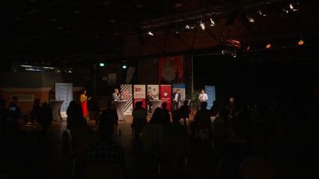Die OB-Kandidat*innen auf dem Podium aus der hinteren Reihe heraus fotografiert Die OB-Kandidat*innen auf dem Podium aus der hinteren Reihe heraus fotografiert