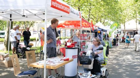 Pavillon mit der Aufschrift: Verbraucherzentrale. Vor dem Stand steht eine Frau mit einem elektrischen Rollator, die Richtung eines Mannes blickt. Dieser steht im Pavillon. 