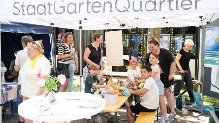 An einem Pavillon steht die Aufschrift "Stadtgartenquartier". Darunter befinden sich mehrere Personen an Stehtischen.