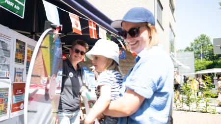 Eine Frau mit Cappy und Sonnenbrille hebt ein Kleinkind zu einer bunten Drehscheibe hoch. Daneben steht eine lächelnde Frau.