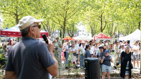 Ein Mann mit Cappy und Sonnenbrille spricht in ein Mikrofon zu einer größeren Menge an Menschen auf einem Platz. Auf diesem stehen auch Pavillons und Bäume. 