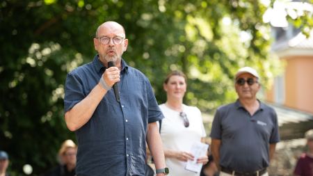 Vor einem Baum steht der Politiker Rainer Schmeltzer und spricht in ein Mikrofon. Schräg dahinter stehen die Bürgermeisterin Martina Förster-Teutenberg und Kenan Küçük.