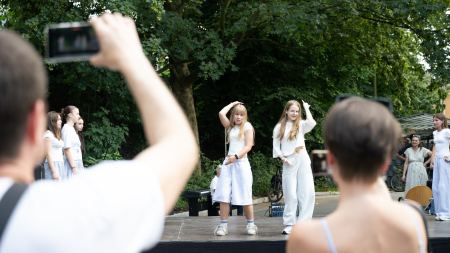 Auf einer Bühne stehen tanzende Mädchen. Davor stehen Menschen, die fotografieren. 