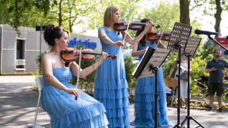 Drei Violinistinnen spielen auf ihrem Instrument. Sie tragen blaue Abendkleider und blicken auf Notenblätter, die auf Notenständern liegen. 