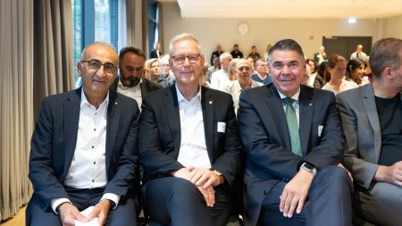 Drei Männer in Anzügen sitzen nebeneinander in der ersten Reihe in einem Saal.