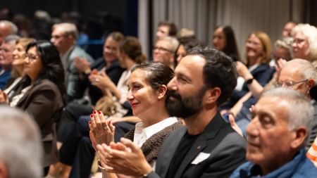 Applaudierendes Publikum sitzt auf Stuhlreihen in einem Saal. 