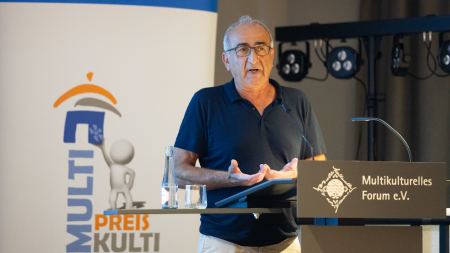 Ein Mann steht an einem Stehpult. Hinter ihm befindet sich ein Banner mit der Aufschrift: Multi-Kulti-Preis.