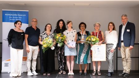 Auf einer Bühne stehen nebeneinander aufgereiht zahlreiche Personen - einige halten Blumensträuße, einen Pokal und einen Bilderrahmen.