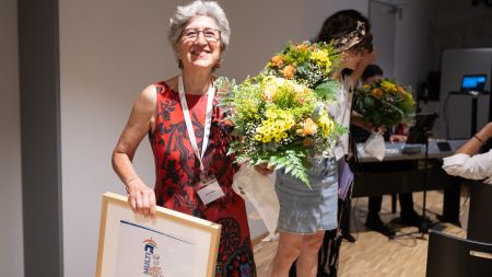 Eine Frau trägt einen Blumenstrauß, eine Urkunde und ein großes Lachen.  