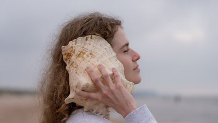 Eine Frau steht am Strand und hält eine Muschel an ihr Ohr. Der Himmel ist bewölkt. Sie trägt einen grauen Pullover und hat lockige dunkelblonde Haare, die ihr bis über die Schultern reichen. 
