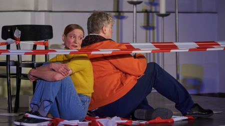 Ein Mann und eine Frau sitzen auf einer Bühne auf dem Boden. Sie sind von rot-weißem Absperrband eingekreist.
