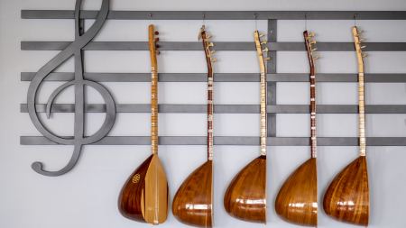 An einer Wand befindet sich ein großflächiger Violinschlüssel mit Notenlinien. Davor hängen hintereinander aufgereiht fünf Bağlama-Langhalslauten.