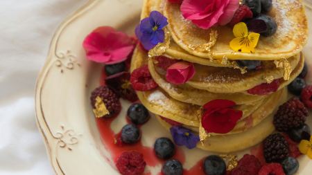 Auf einem verzierten Teller ist ein Stapel Pancakes mit Blüten und Blattgold angerichtet. 