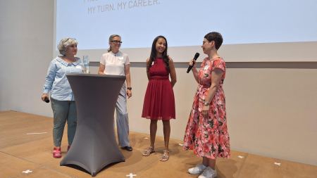 Vier Frauen stehen nebeneinander vor einer Leinwand. Eine Frau hält ein Mikrofon vor ihren Mund.