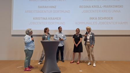 Fünf Personen stehen auf einer Bühne. Eine Frau hält ein Mikrofon in ihren Händen.