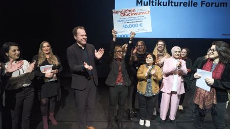 Frau hält freudig Preisscheck in die Höhe, andere stehen gemeinsam mit ihr auf der Bühne