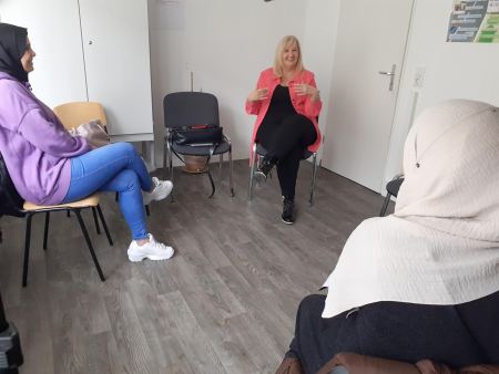 Drei Frauen sitzen in einem Stuhlkreis. Zwei davon tragen ein Kopftuch und blicken die dritte Frau an. Diese gestikuliert mit den Händen.