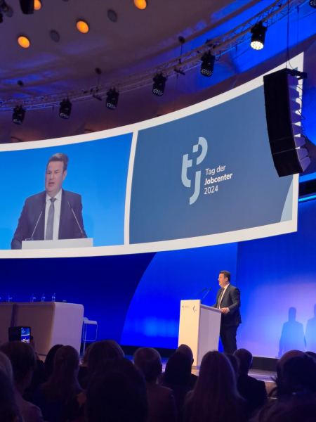 Bundesarbeitsminister Hubertus Heil steht auf einer großen Bühne an einem Rednerpult und hält eine Rede. Im Hintergrund ist eine hohe, blau beleuchtete Wand zu sehen auf die die Live-Aufnahme Heils abgespielt wird.