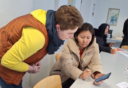 Bewerbungstraining im Projekt MY TURN. MY CAREER. Eine stehende Frau lehnt sich über eine sitzende Frau, die ein Handy festhält. Beide schauen auf den Handybildschirm.