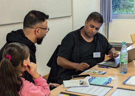 Auf einer Tischplatte liegen zahlreiche Dokumente. Dahinter sitzen ein Mädchen, ein Mann mit Brille und Bart und ein weiterer Mann mit Namensschild auf dem T-Shirt, den die anderen anblicken.