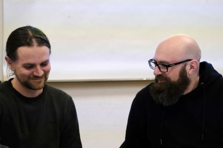 Zwei Männer sitzen nebeneinander, beide tragen schwarze Pullover: Sebastian Rose (links) hat braune Haare und einen Bart und blickt zu Boden. Sascha Schiessl (rechts) hat einen braunen Bart, eine Glatze und eine Brille. Er schaut zum linken Bildrand.