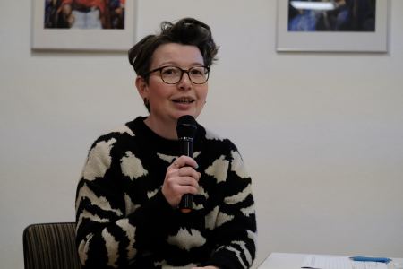 Eine Frau mit kurzen Haaren und einer Brille trägt einen Wollpulli und hält sich mit beiden Händen ein Mikrofon vor den Mund. Sie sitzt auf einem Stuhl vor einer Wand mit Bildern. 