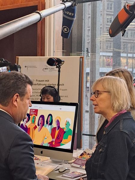 Bundesarbeitsminister Hubertus Heil unterhält sich an einem Messestand mit einer Frau. Am Messestand ist ein Laptop zu sehen, auf dem die Website des Bundesprogramms My Turn aufgerufen ist.