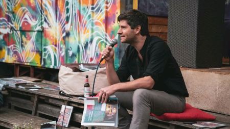 Alexander Estis sitzt während einer Veranstaltung neben bunt besprayter Wand auf einer Bank und spricht in ein Mikrofon
