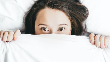 Frau blickt mit weit geöffneten Augen unter weißer Bettdecke hervor