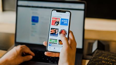 Person hält Smartphone mit einer Hand und bedient Laptop mit anderer Hand