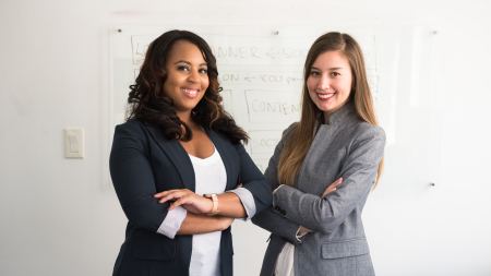 Zwei Frauen in Business-Outfits stehen lächelnd und mit verschränkten Armen vor einer Whiteboard