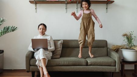 Mutter arbeitet mit Laptop auf Couch, auf der ihr KInd hüpft