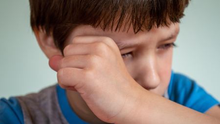 Ein Kind mit kurzen braunen Haaren hält sich die geschlossene Hand vor das Gesicht. 