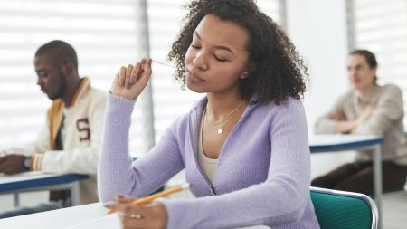 Schwarze Frau mit lila Oberteil in Prüfungssituation in Klassenraum mit Stift in der Hand. Im Hintergrund sieht man zwei weitere Prüflinge