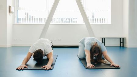 Zwei Personen dehnen sich auf zwei Sportmatten in einem hellen Raum.
